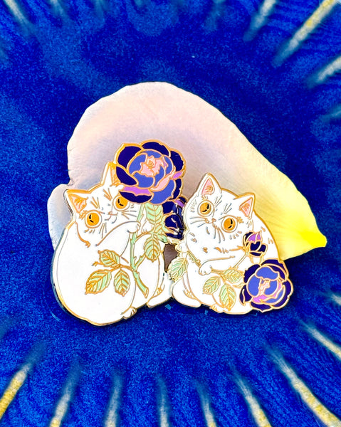 Two cat-shaped pins with floral details on a blue shell background